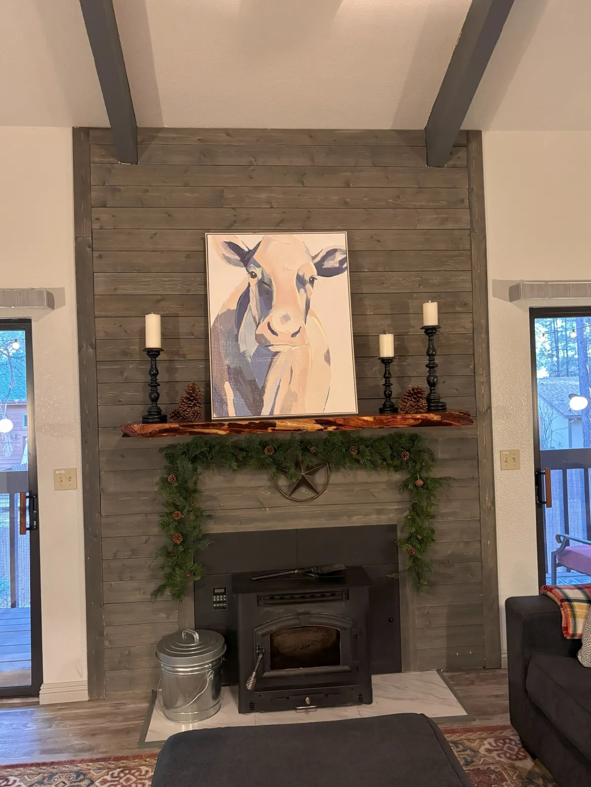 The image features a cozy great room with a fireplace as the focal point, flanked by a tasteful cow painting and wooden shelf adorned with candles and greenery. The scene is warm and inviting, with comfortable furnishings arranged for group gatherings and intimate relaxation. Large windows likely fill the space with natural light, contributing to an open and airy feel.