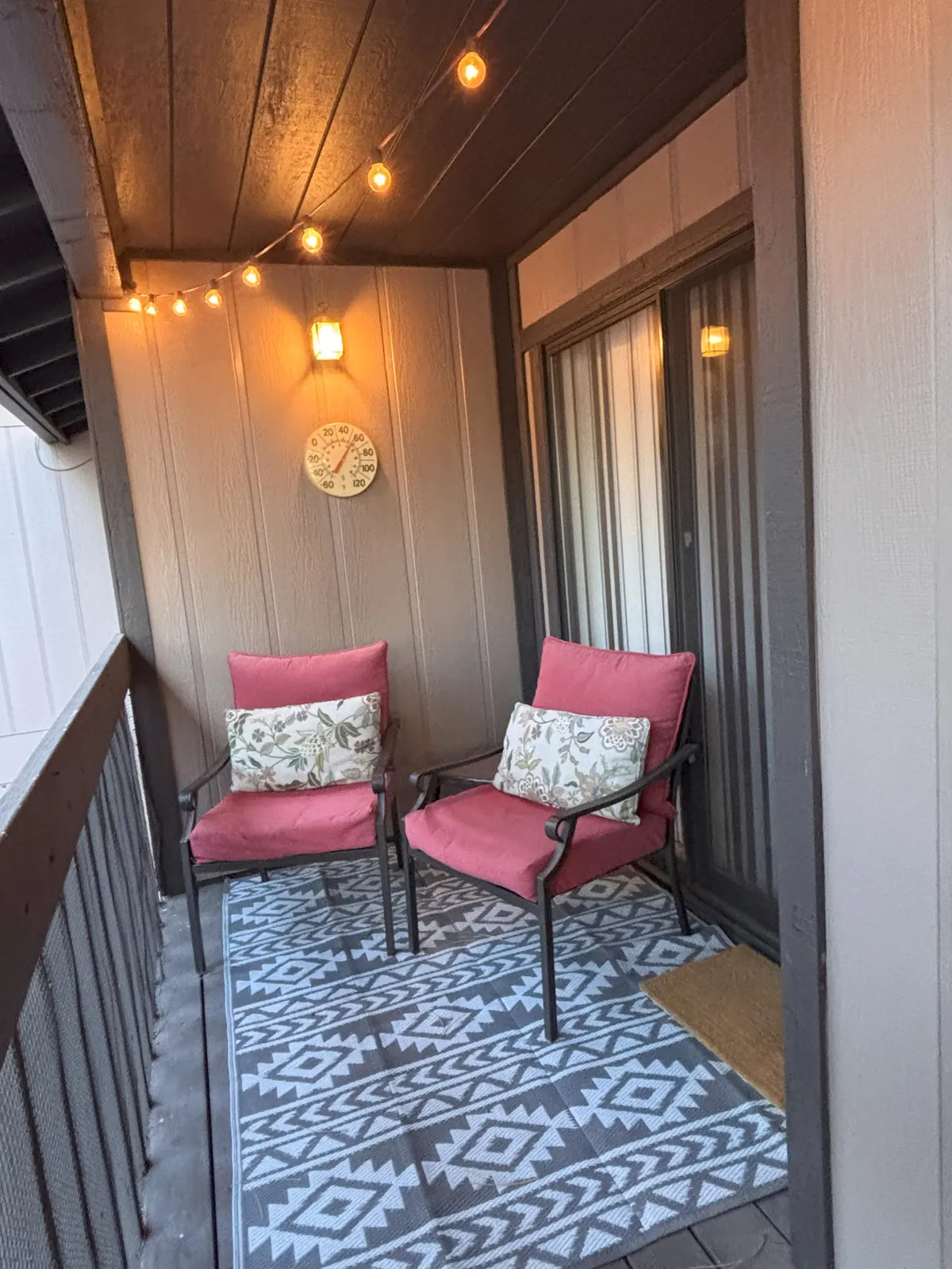 A cozy exterior patio scene with two comfortable chairs, a decorative rug, string lights, and a wall clock. The intimate arrangement suggests evenings spent in conversation or quiet relaxation, surrounded by the ambiance of nature.