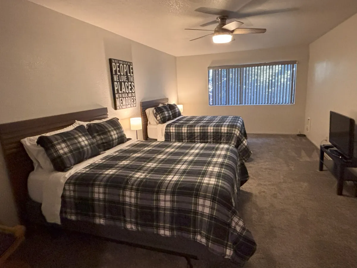A bedroom featuring two inviting plaid beds, a large window with blinds letting in natural light, and a wall art piece with text. The space feels bright, comfortable, and ideal for families or friends sharing a room.